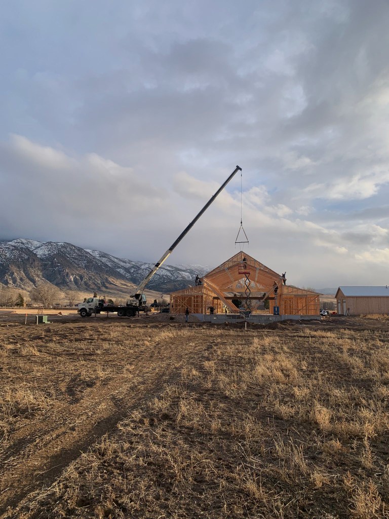 Boom truck crane on a rural construction site. Barndominium trusses are being lifted from a spreader bar and secured by a framing crew.