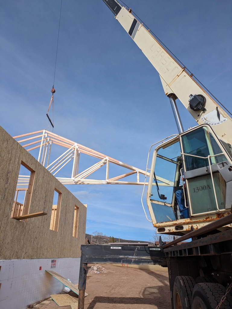 View of a crane lifting large trusses onto a building. Angle shows the operating cab and part of the boom.
